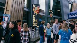 Lots of people stood outside the Students' Union building, there are Sports Fair flags