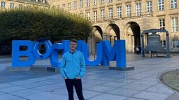 A male student stands in front of a 3D 'Bochum' sign and smiles at the camera. It is a sunny day and there is a large building in the background.