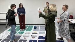 A group of students play the life size boardgame, one is holding a card and reading aloud, whilst everyone stands on the board in their own space.