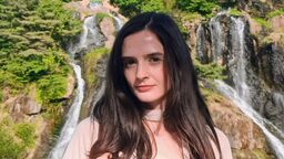 A young female stands in front of a waterfall and looks towards the camera.
