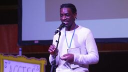Sipho on stage. He holds a microphone and a notebook, and wears a white t-shirt.