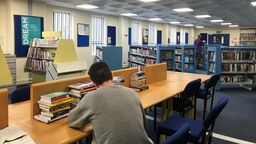 A library with a student sat at a desk and several shelves of books