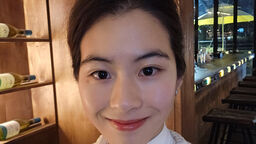 A female student stands in a restaurant and smiles at the camera. The lighting is low and behind them are bottles of wine on shelves.