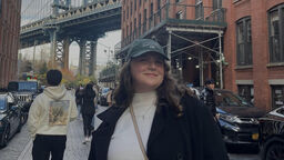 A female student stands in the foreground of an urban street in the USA, wearing a baseball cap and smiling at the camera. Behind them is a bridge in the background.
