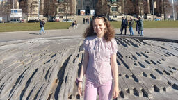 A female student stands outdoors with a large historical building and blue sky behind them. They are smiling at the camera.