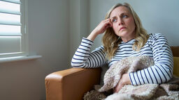 A woman sitting on a sofa, covered in a blanket, looking tired and fed up.
