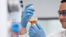 A lab worker is holding up a bottle with an orange liquid inside.