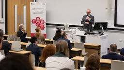A man in a grey suit is speaking into a microphone to people in a lecture theatre