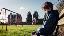 Child sitting on bench in a park during the daytime. They look sad.
