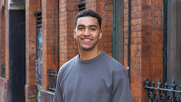 Architecture graduate smiling outside Manchester red brick building in grey jumper and jeans 