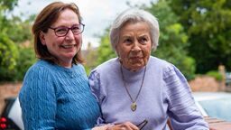 A middle-aged woman smiles at the camera while helping an older woman to walk by guiding her arm.
