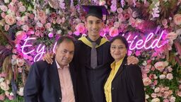 Student Vayam Lahoti wearing graduation cap and gown, with a man and a woman, stood in front of fake flowers and a neon sign
