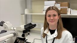 A person smiling in a lab coat sitting by a microscope