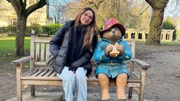 A sunny autumnal/wintery day; a woman wrapped up in a black puffer jacket, scarf, gloves and light jeans sits smiling on a bench in a park leaning in towards a Paddington Bear statue. The statue is eating the classic marmalade sandwich, which the character is known for. Paddington is wearing his classic blue toggle coat, and red hat. Behind them on the bench, there are wintry trees missing their leaves and a few obscure buildings.